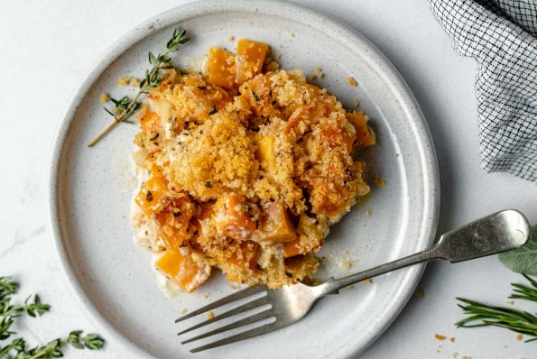 Butternut squash au gratin shown on a small plate with a fork. The plate sits atop a light blue surface, next to some fresh herbs & a checkered linen napkin.