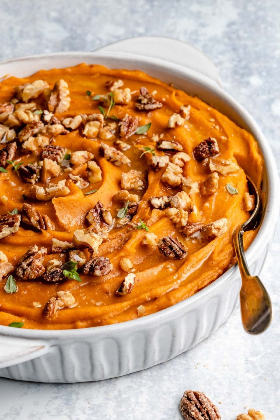 Easy Sweet Potato Casserole in a white serving dish sprinkled with fresh herbs and candied nuts with a silver spoon nestled into the dish. The baking dish rests on a flat marbled gray surface.