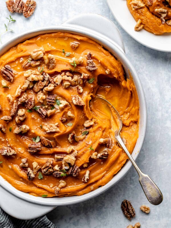 Easy Sweet Potato Casserole in a white serving dish sprinkled with fresh herbs and candied nuts with a silver spoon nestled into the dish. The baking dish rests on a flat marbled gray surface next to a gray and white stripe linen napkin, a plate of sweet potato casserole dished up and candied nuts and herbs spread around.