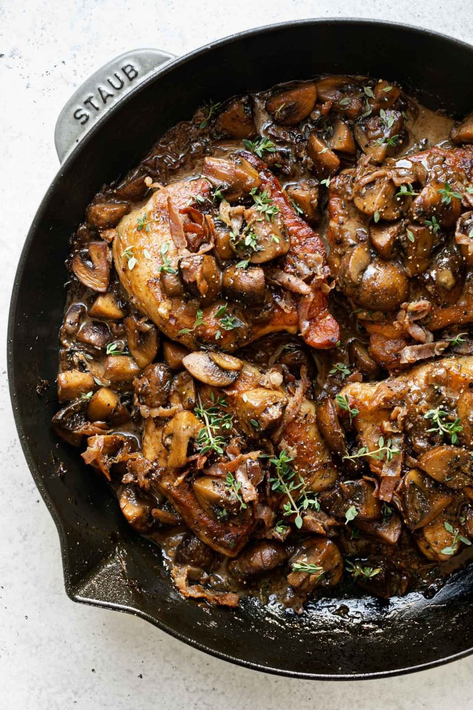 Pork marsala with mushrooms & creamy marsala sauce in large gray Staub skillet. The pork chops are topped with caramelized mushrooms, crispy prosciutto & fresh thyme. The skillet sits atop a white surface.
