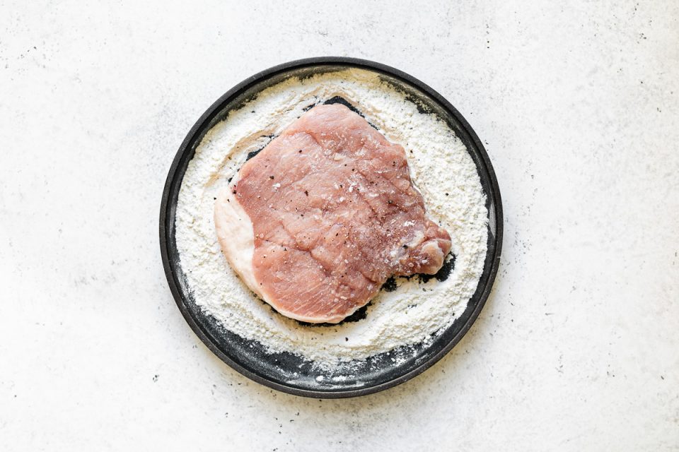 Dredging pork chop in flour on a small black plate. The plate sits atop a white surface.