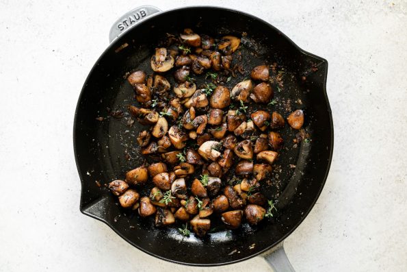Caramelized mushrooms garnished with fresh thyme in a large gray Staub skillet. The skillet sits atop a white surface.