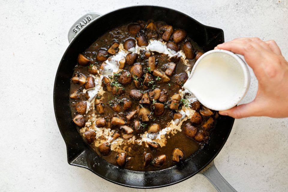 Woman's hand pouring heavy cream into skillet with mushrooms & marsala sauce. The skillet sits atop a white surface.