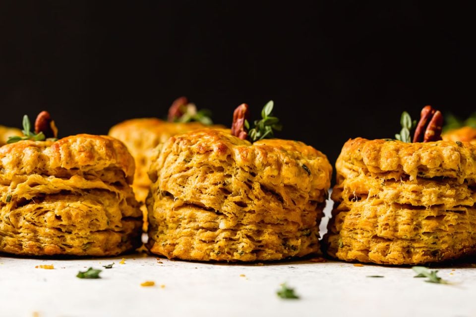 Side angle of a row of pumpkin biscuits. The biscuits have a pecan & fresh herbs inserted in their tops, making the biscuits look like little pumpkins.