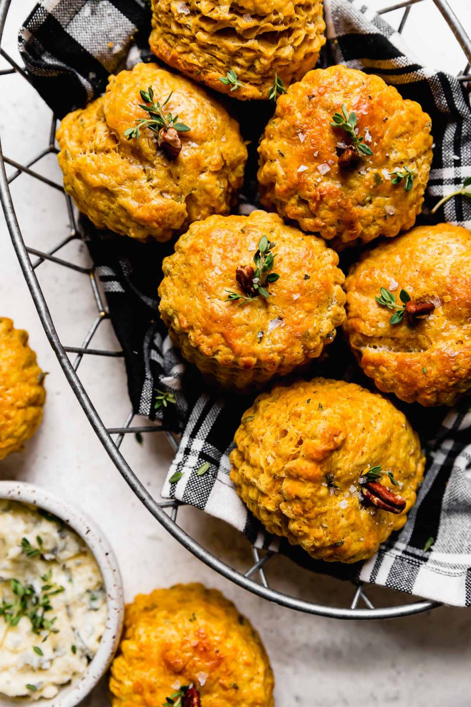 Pumpkin biscuits, decorated like pumpkins with a pecan & fresh herbs inserted into the tops, shown in a wire basket lined with a black plaid napkin. The basket sits atop a white surface next to a small ceramic bowl with garlic herb butter.
