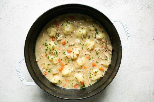 Italian chicken & gnocchi dumplings soup shown in white Staub dutch oven on a white surface. The soup is garnished with finely chopped fresh chives.