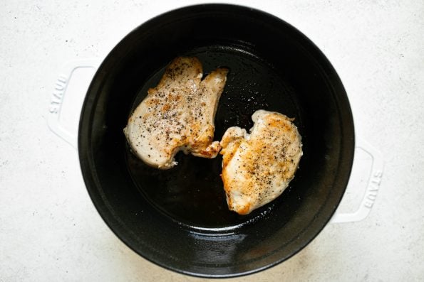 2 browned chicken breasts shown in a white Staub Dutch oven placed on a white surface.