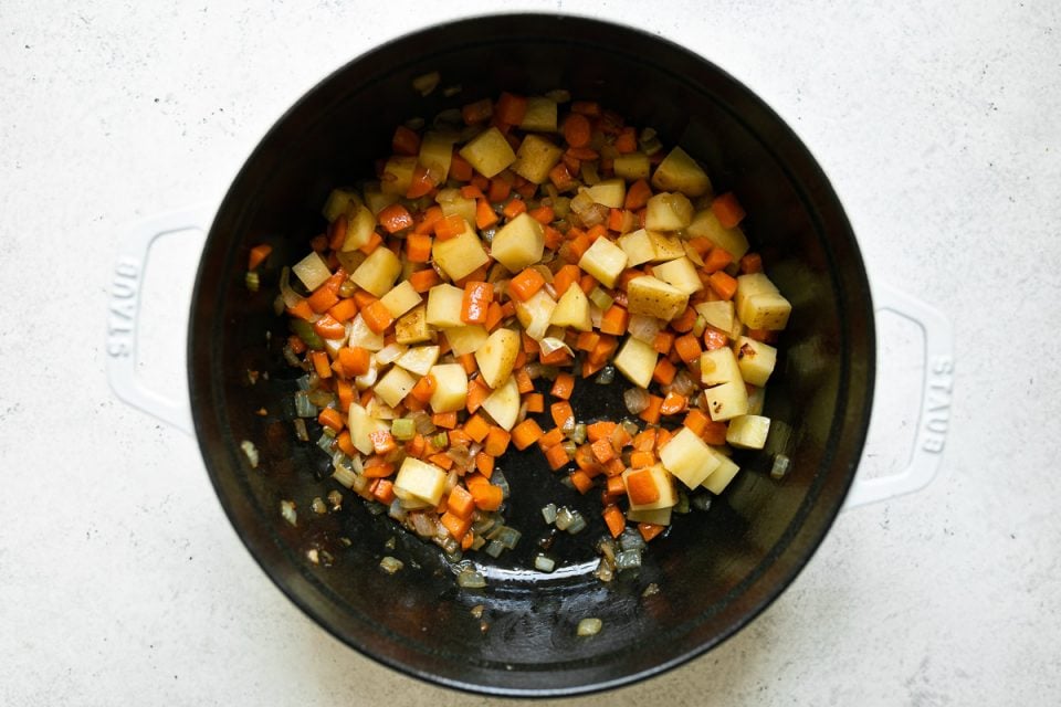 Sauteed veggies (onions, celery, carrot & potatoes) shown in a white Staub Dutch oven placed on a white surface.