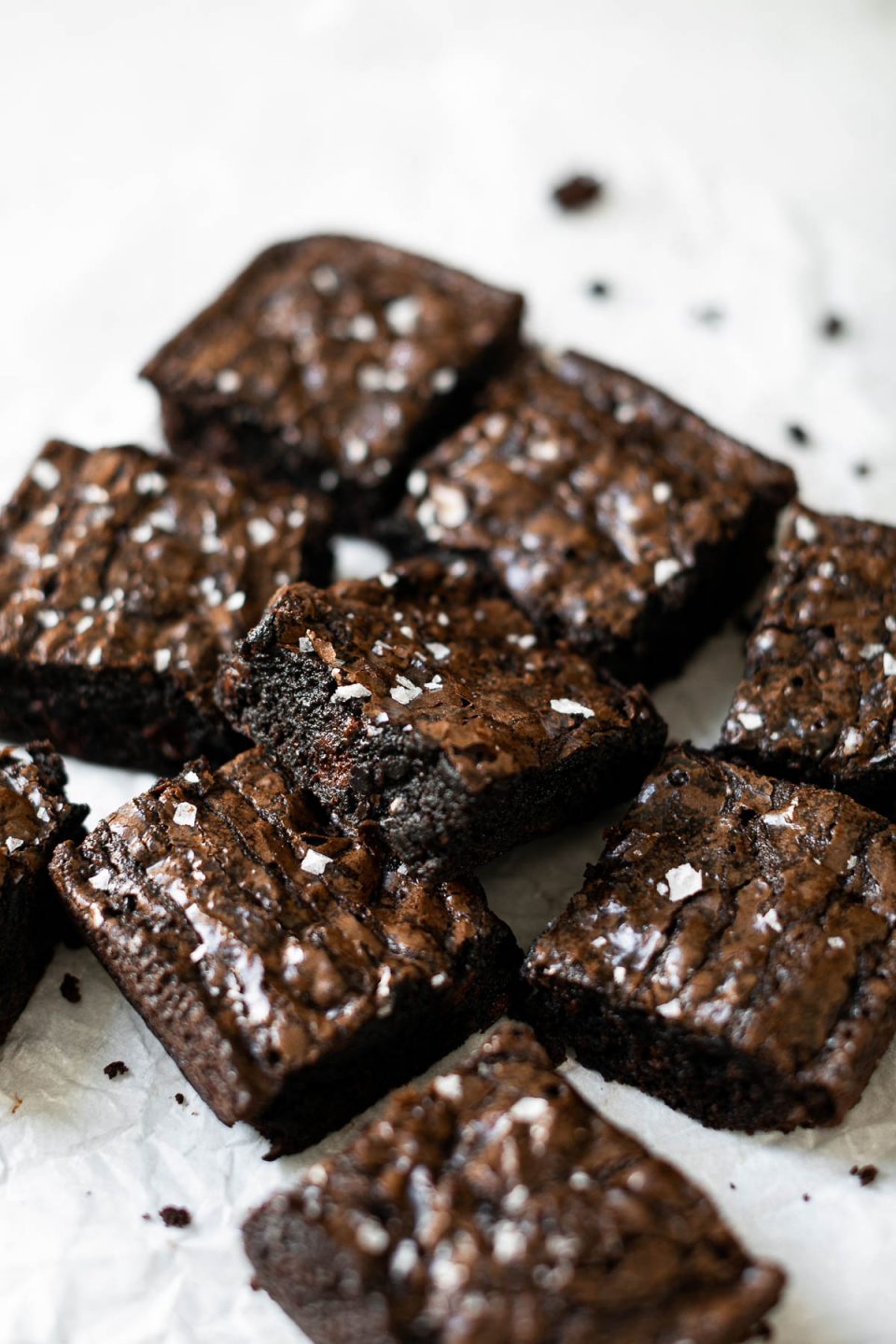 Multiple brownies cut into squares arranged on a piece of white crumpled parchment paper & topped with flaky sea salt.