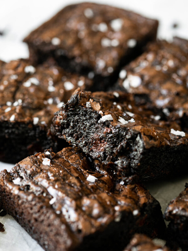 A stack of brownies made with box brownie hacks and Ghirardelli brownie box recipe cut into squares arranged on a piece of white crumpled parchment paper & topped with flaky sea salt.