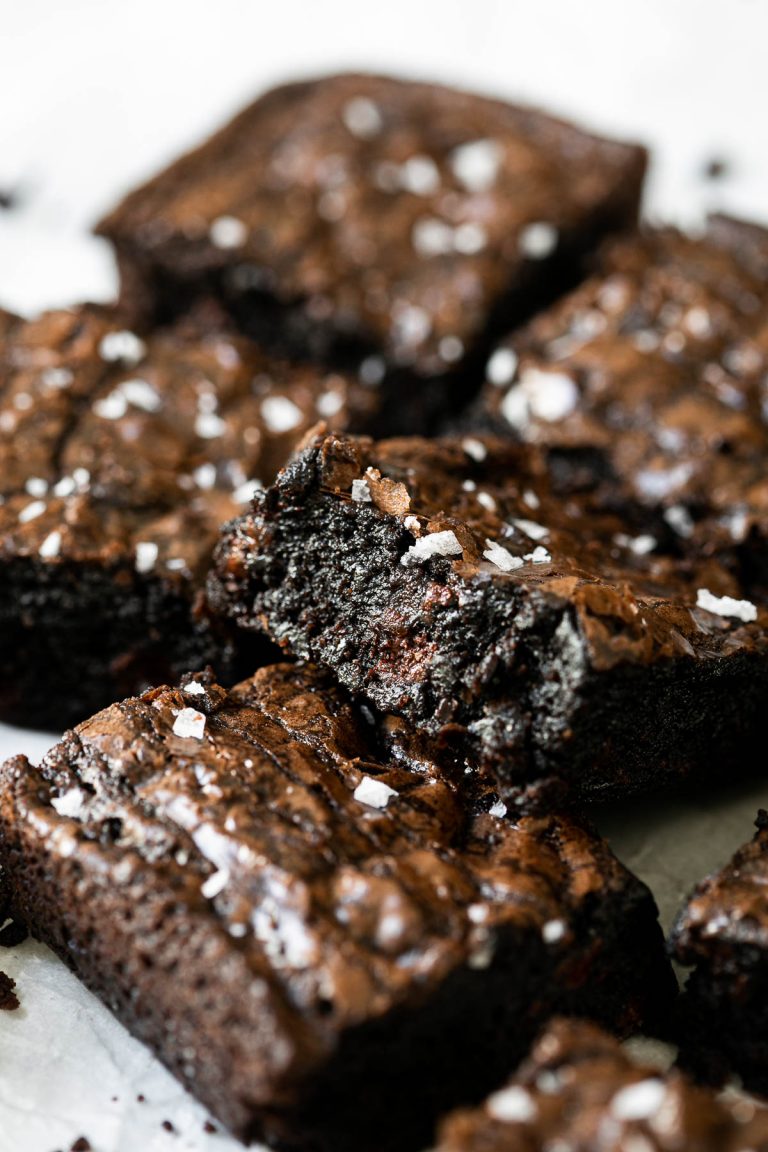 A stack of brownies made with box brownie hacks and Ghirardelli brownie box recipe cut into squares arranged on a piece of white crumpled parchment paper & topped with flaky sea salt.