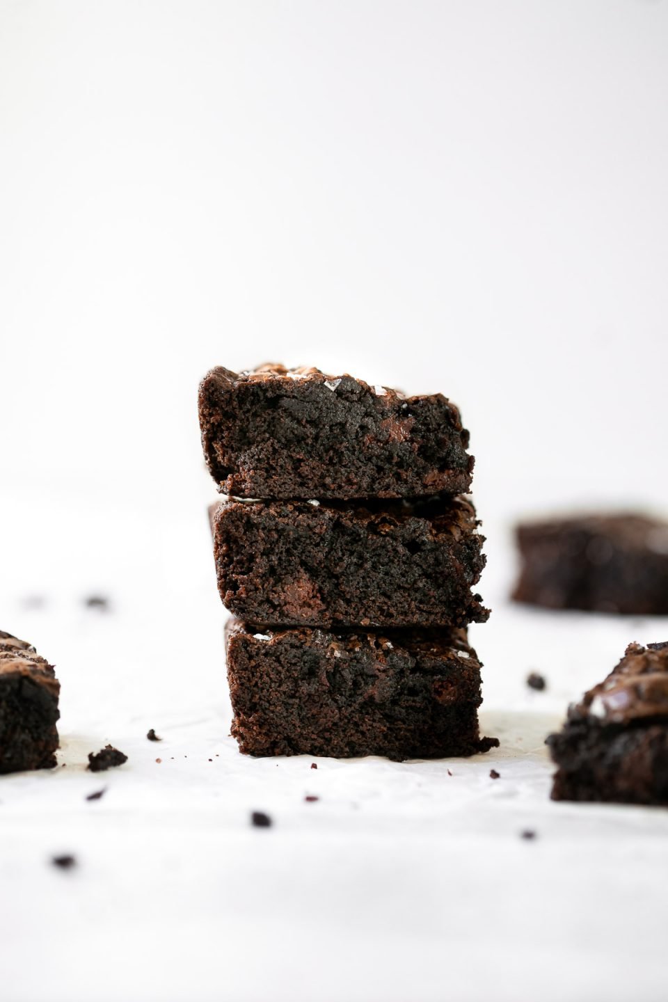 A side angle shot of a stack of 3 fudgy lazy girl brownies topped with flaky sea salt atop a crumpled piece of parchment paper. Single set on the same surface brownies surround the stack.