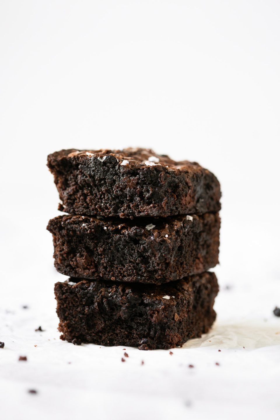 A side angle shot of a stack of 3 fudgy lazy girl brownies made with steps for how to make box brownies better and a Ghriradelli box brownie recipe. The brownies have been topped with flaky sea salt atop a crumpled piece of parchment paper.