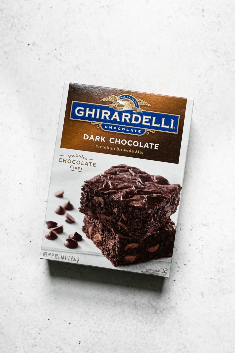 A overhead shot of Ghirardelli Dark Chocolate Premium Brownie Mix which is used in the recipe to make box brownie recipe rests with front of box facing up on a white surface.