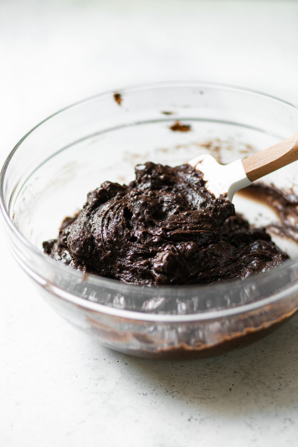 Brownie batter made with boxed brownie mix and added box brownie hacks prepped in a clear glass mixing bowl. A white spatula with a wooden handle is stuck inside the brownie batter. The mixing bowls sits atop a white surface.