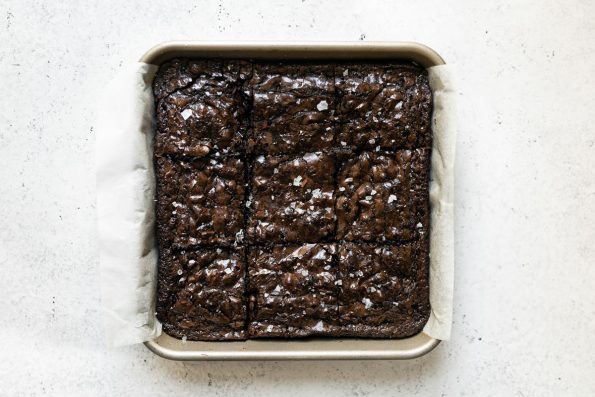Lazy Girl Brownies made from box brownies baked in a parchment-lined baking pan. The brownies have been cut yielding 9 brownie squares. The pan sits atop a white surface.