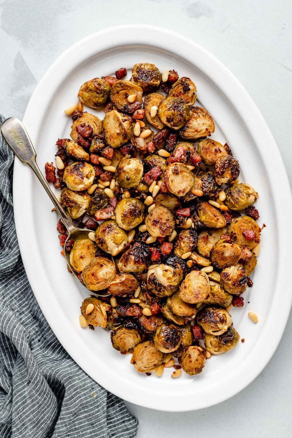 Maple roasted brussels sprouts on a large white serving dish, with pancetta & pine nuts. The white plate sits atop a light blue surface, next to a striped blue linen napkin.