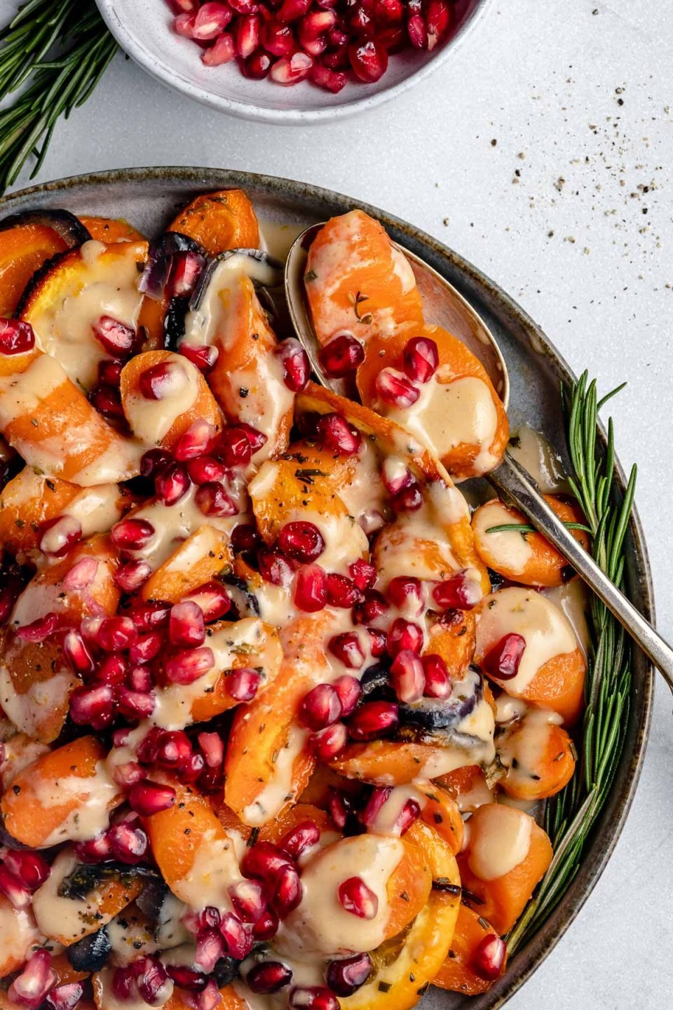Perfectly roasted maple glazed carrots, red onions, and orange slices in a large gray serving platter drizzled with an orange tahini sauce and sprinkled with pomegranate arils and with a sprig of fresh rosemary on the side. A silver spoon is nestled into the dish and rests on the platter. The platter sits on top of a light gray surface with a light gray bowl of pomegranate arils in the background and another sprig of fresh rosemary and cracked black pepper surrounding the bowl.