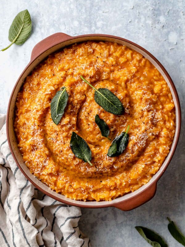 Creamy pumpkin risotto shown in a large terra cotta serving dish. The risotto is topped with crispy fried sage leaves & finely grated parmesan. The dish sits atop a light blue surface, next to a striped linen napkin & some fresh sage leaves.