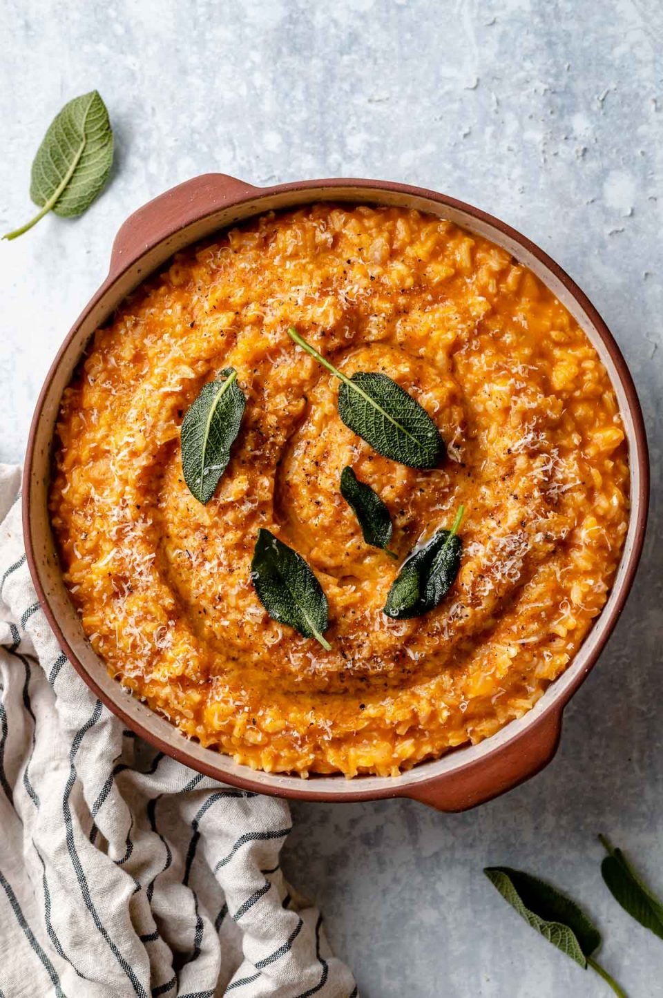 Creamy pumpkin risotto shown in a large terra cotta serving dish. The risotto is topped with crispy fried sage leaves & finely grated parmesan. The dish sits atop a light blue surface, next to a striped linen napkin & some fresh sage leaves.