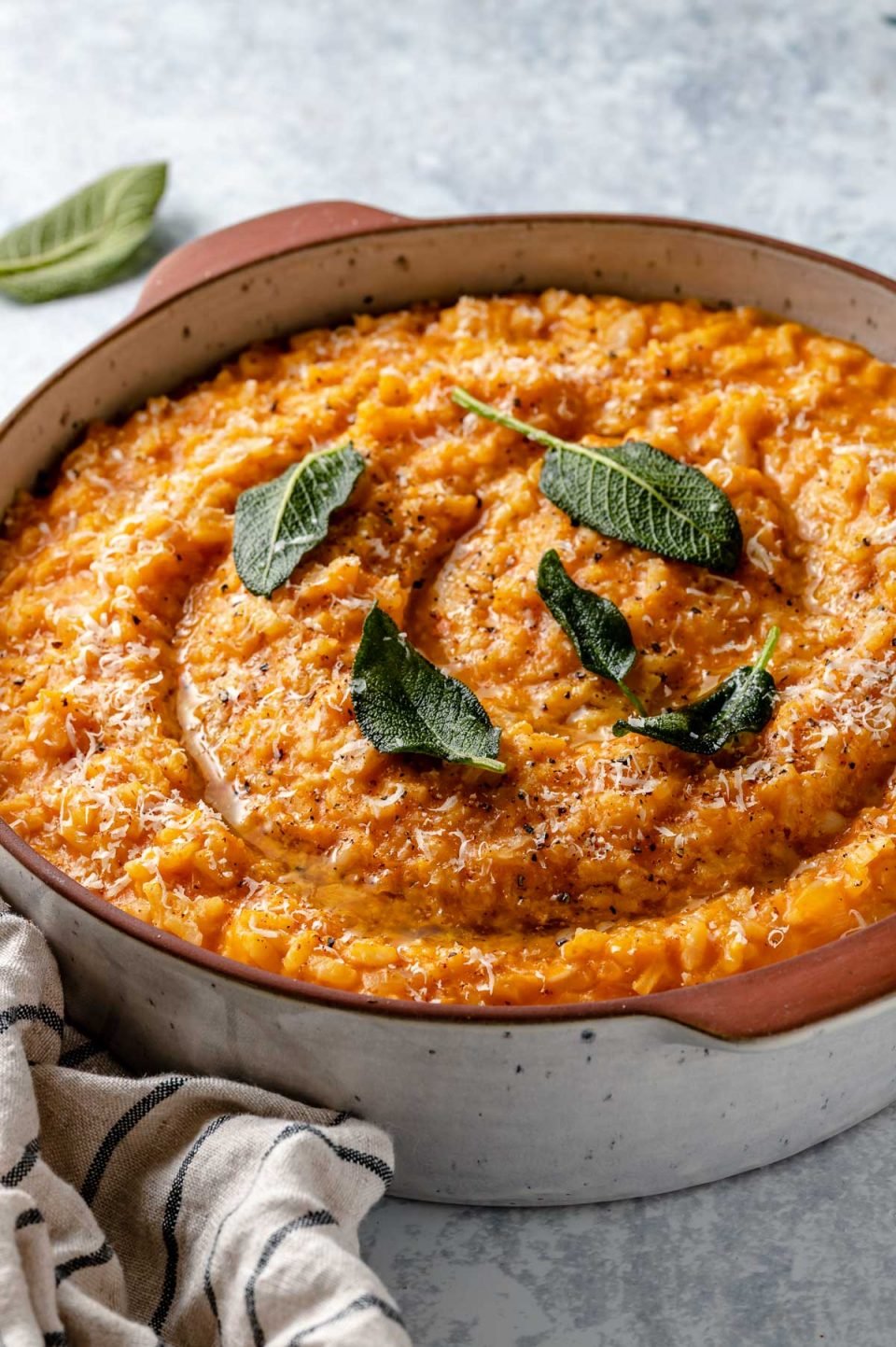 Side angle of creamy pumpkin risotto shown in a large terra cotta serving dish. The risotto is topped with crispy fried sage leaves & finely grated parmesan. The dish sits atop a light blue surface, next to a striped linen napkin & some fresh sage leaves.