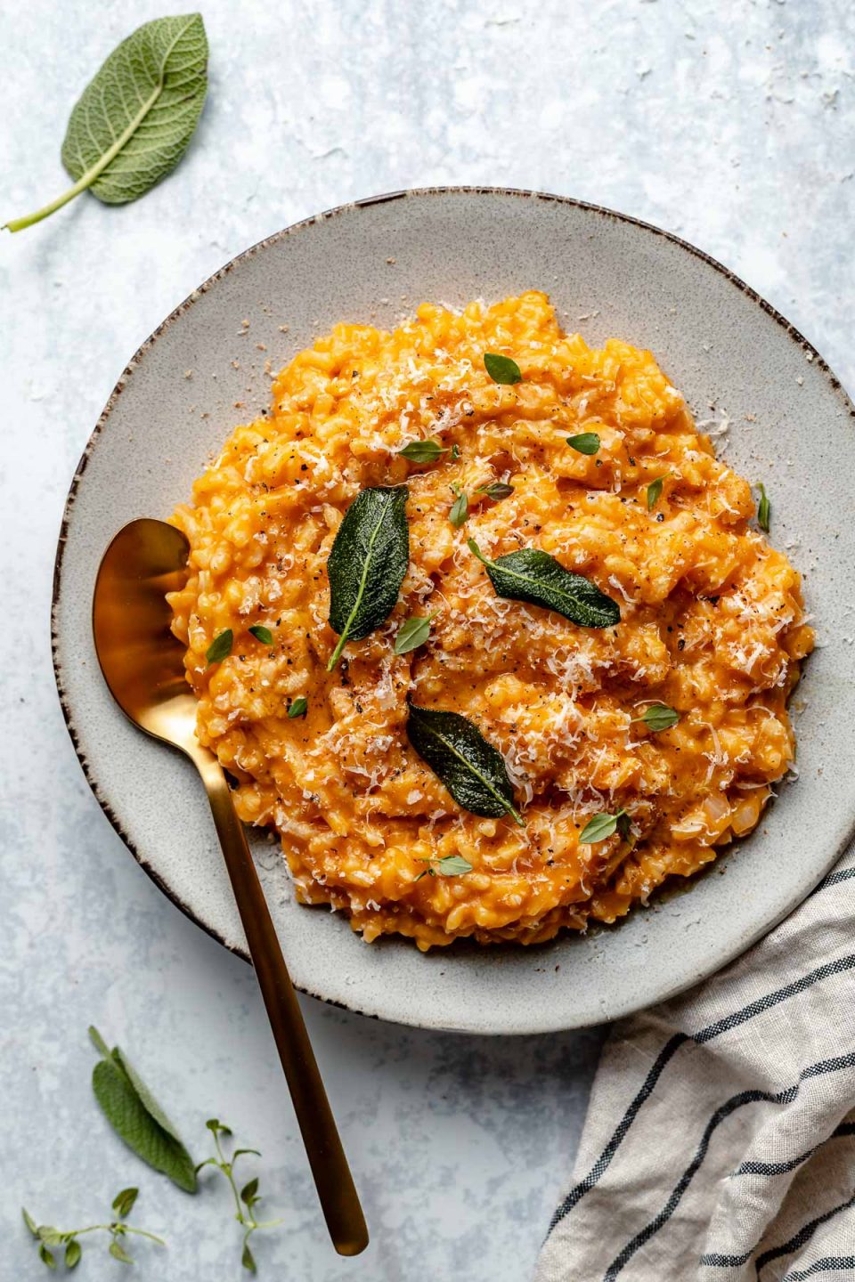 Creamy pumpkin risotto in a large serving dish. The risotto is topped with freshly grated parmesan & crispy fried sage leaves. There is a gold spoon nestled into the risotto. the bowl sits atop a light blue surface, next to a striped linen napkin & fresh sage leaves.