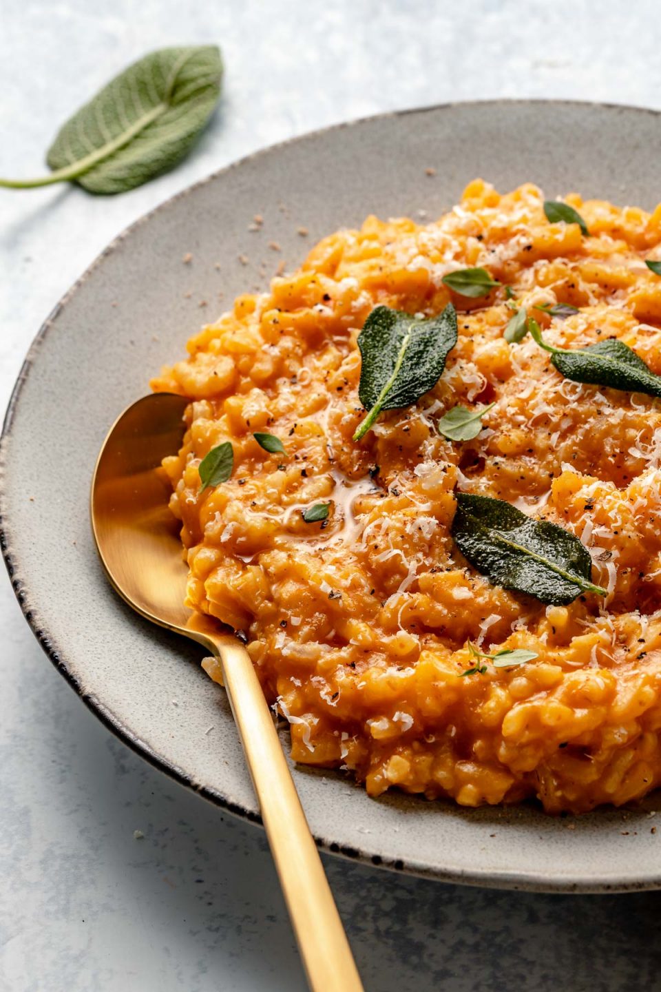 Side angle of creamy pumpkin risotto in a large serving dish. The risotto is topped with freshly grated parmesan & crispy fried sage leaves. There is a gold spoon nestled into the risotto. the bowl sits atop a light blue surface, with fresh sage leaves in the background.