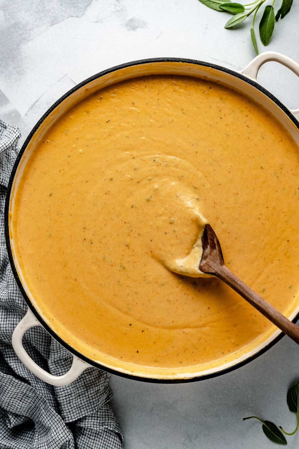 Creamy Pumpkin Mornay sauce in a large white pan with a wooden spoon stirring it. The pan sits atop a light blue surface, next to a checkered linen & fresh sage leaves.