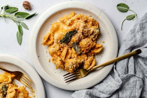 Creamy pumpkin macaroni & cheese plated on white ceramic plates. The plates sit atop a light blue surface, next to a checkered linen & fresh sage leaves.
