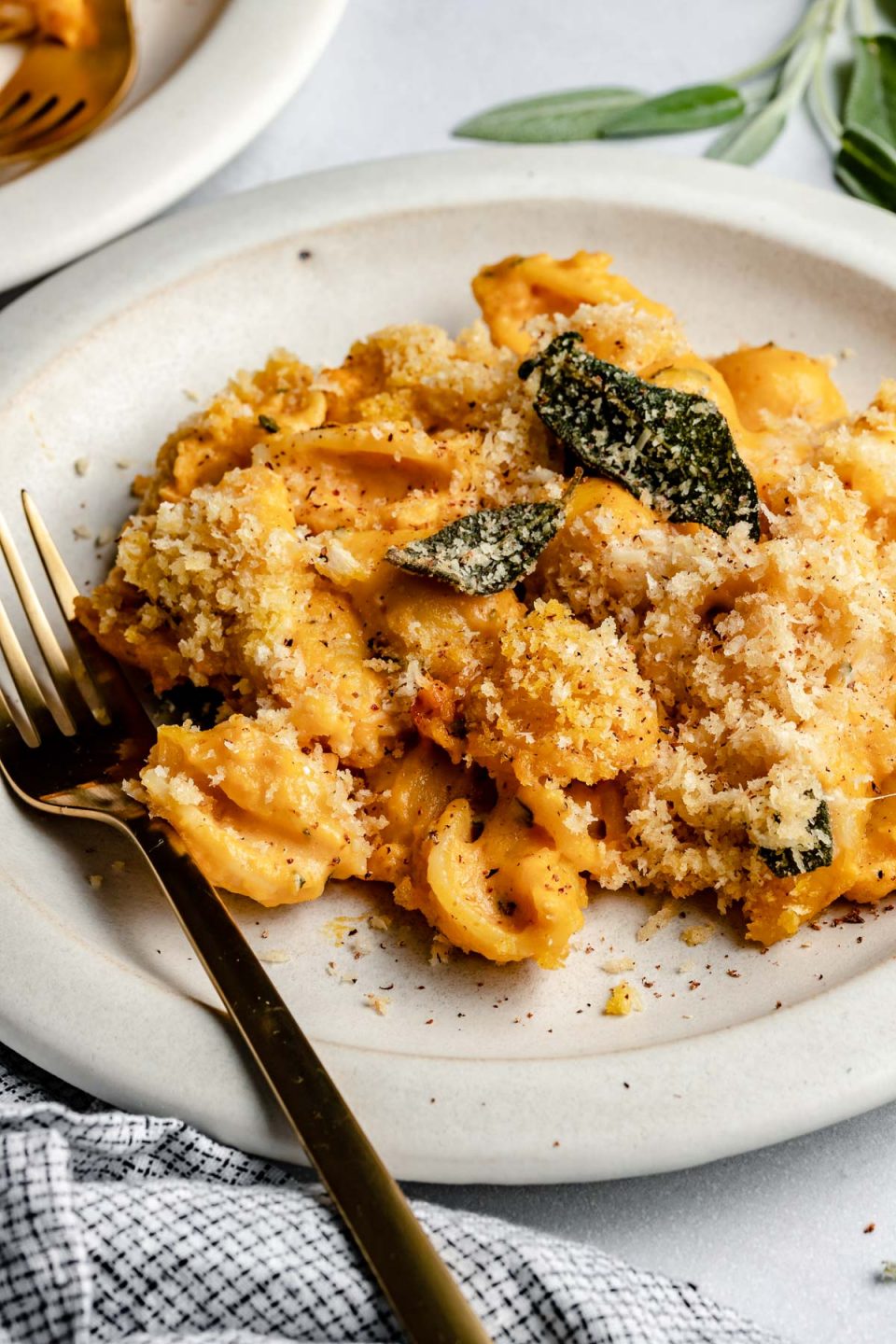 Side angle of creamy pumpkin macaroni, topped with brown butter sage breadcrumbs. The macaroni is on a ceramic white plate, next to a checkered linen & fresh sage leaves.