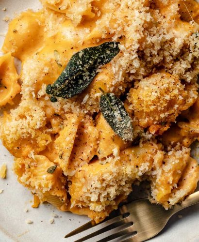 Creamy pumpkin mac & cheese shown on a white ceramic plate, topped with brown butter breadcrumbs & crispy sage leaves.
