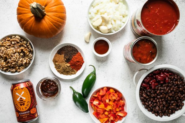 Pumpkin chili ingredients arranged on a white surface: a bowl of walnuts, small sugar pumpkin, Ichabod Pumpkin Ale, jar of chipotle peppers, plate of chili spices, bowl of diced onions & garlic cloves, 2 jalapeno peppers, bowl of diced peppers, black beans & kidney beans in small white colander, 2 cans of crushed tomatoes.