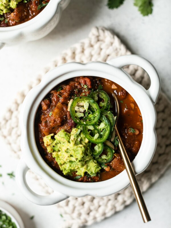 A white, pumpkin-shaped bowl of vegan pumpkin chili, sits atop a white crocheted pot holder. A gold spoon sits in the chili, which is also topped with guacamole & sliced jalapeno. A second bowl of chili peeks into the top of the frame, along with a few cilantro leaves.