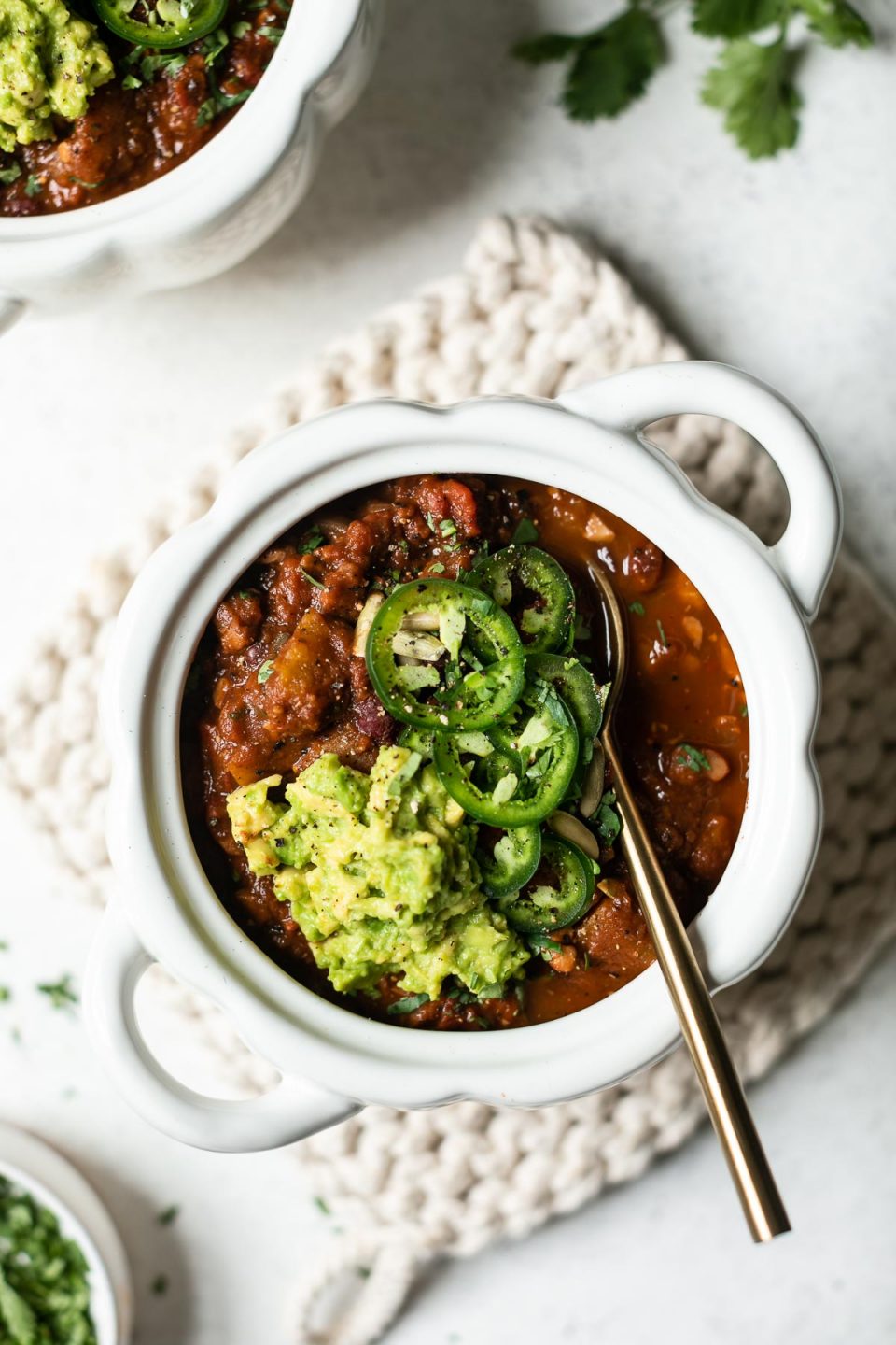 A white, pumpkin-shaped bowl of vegan pumpkin chili, sits atop a white crocheted pot holder. A gold spoon sits in the chili, which is also topped with guacamole & sliced jalapeno. A second bowl of chili peeks into the top of the frame, along with a few cilantro leaves.
