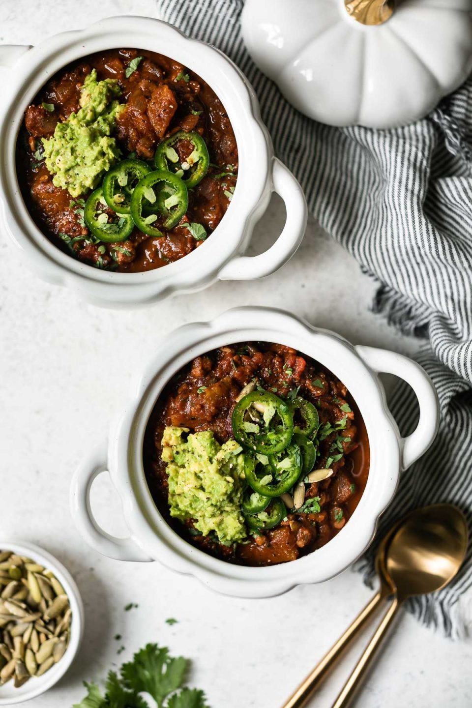 Vegan pumpkin chili shown in 2 small white pumpkin-shaped bowls. The chili is topped with guacamole & sliced jalapeno. The bowls sit atop a white backdrop with a striped linen napkin, 2 gold spoons, a small bowl of pepita pumpkin seeds, & a few cilantro leaves.