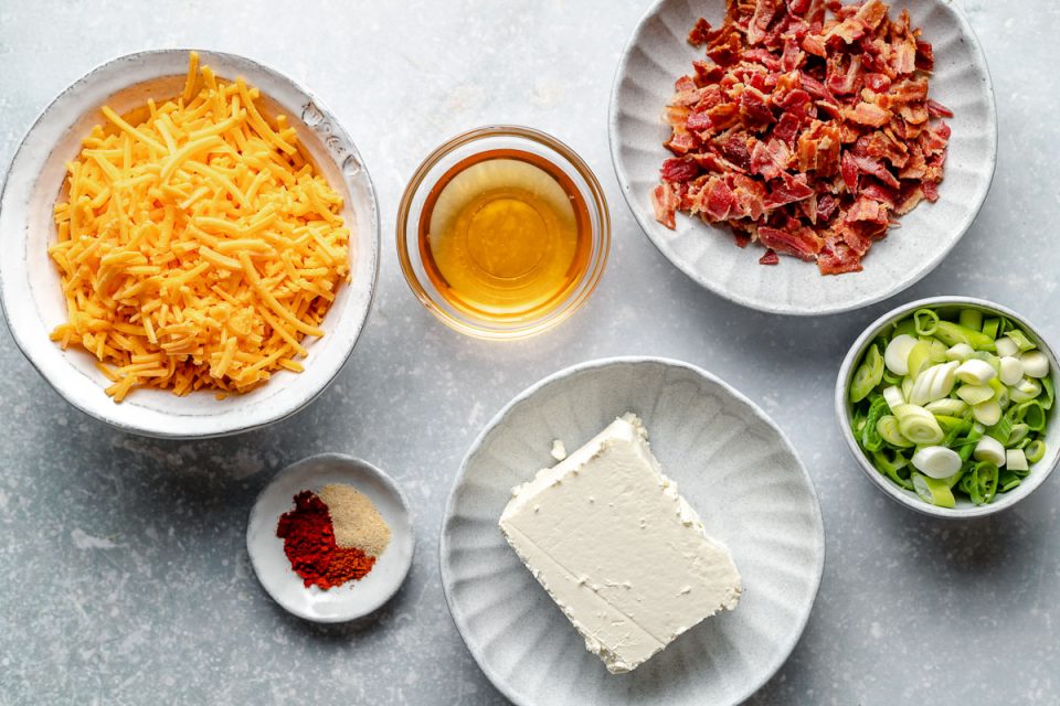 Bacon Cheddar Cheese Ball ingredients arranged on a blue-gray textured surface: cooked & crumbled thick-cut bacon, shredded sharp cheddar, beer, green onions, garlic powder, smoked paprika, cayenne pepper.