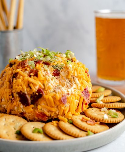 A side angle shot of a Bacon Cheddar Cheese ball sits atop a gray ceramic platter surrounded by a ring of Ritz crackers that have bits of green onion, bacon and shredded cheddar cheese sprinkled on top. The platter rests atop a gray-blue textured surface. A scoop of the cheese ball has been removed. A pint of amber colored beer and a silver container filled with breadstick crackers sits in the background out of focus.
