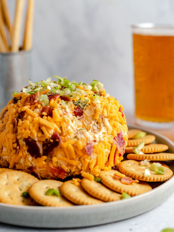 A side angle shot of a Bacon Cheddar Cheese ball sits atop a gray ceramic platter surrounded by a ring of Ritz crackers that have bits of green onion, bacon and shredded cheddar cheese sprinkled on top. The platter rests atop a gray-blue textured surface. A scoop of the cheese ball has been removed. A pint of amber colored beer and a silver container filled with breadstick crackers sits in the background out of focus.