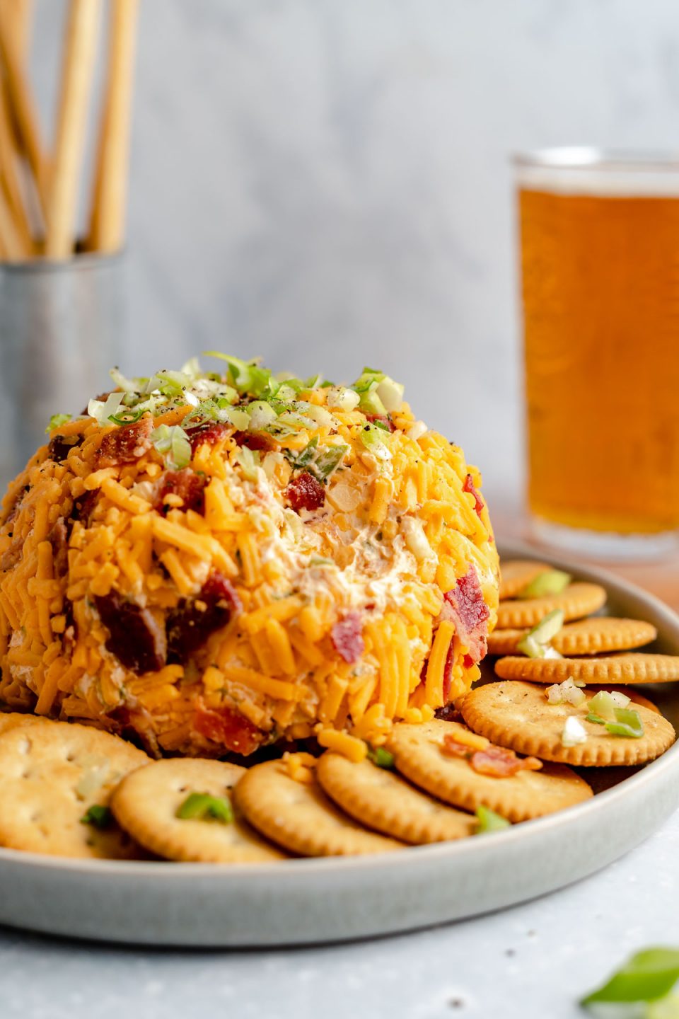 A side angle shot of a Bacon Cheddar Cheese ball sits atop a gray ceramic platter surrounded by a ring of Ritz crackers that have bits of green onion, bacon and shredded cheddar cheese sprinkled on top. The platter rests atop a gray-blue textured surface. A scoop of the cheese ball has been removed. A pint of amber colored beer and a silver container filled with breadstick crackers sits in the background out of focus.
