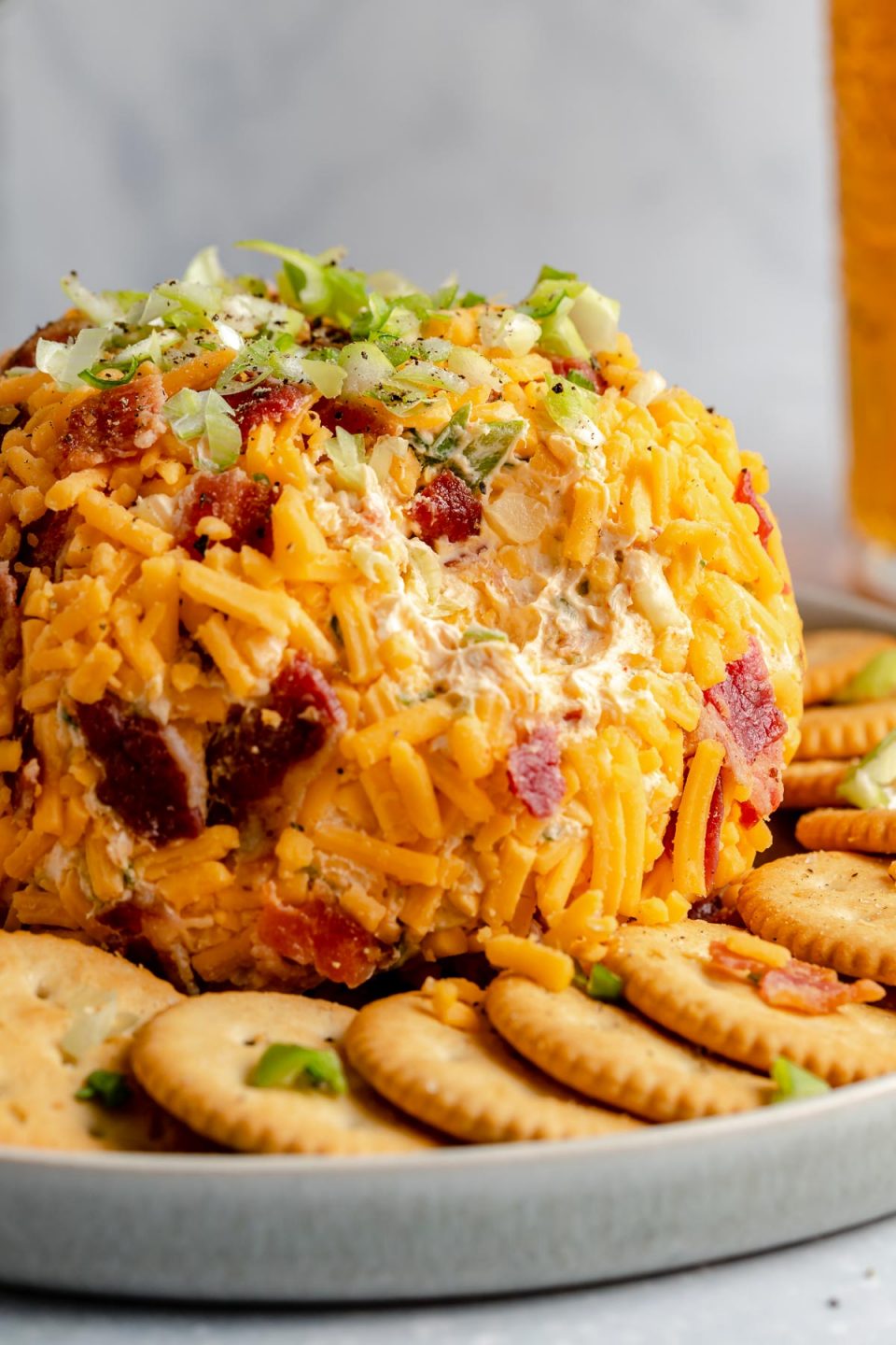 A side angle shot of a Bacon Cheddar Cheese ball sits atop a gray ceramic platter surrounded by a ring of Ritz crackers. The platter rests atop a gray-blue textured surface. A scoop of the cheese ball has been removed and a pint of beer sits in the background out of focus.
