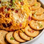 A Bacon Cheddar Cheese ball sits atop a gray ceramic platter surrounded by a ring of Ritz crackers. The platter rests atop a gray-blue textured surface with a single cracker sitting on the surface next to the platter with a scoop of cheese ball atop.