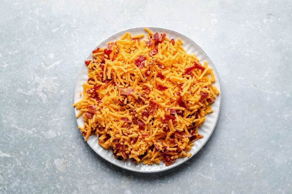 Crumbled cooked bacon and fresh shredded cheddar cheese mixed together for coating a cheese ball sit atop a white ceramic plate. The plate rests atop a blue-gray textured surface.