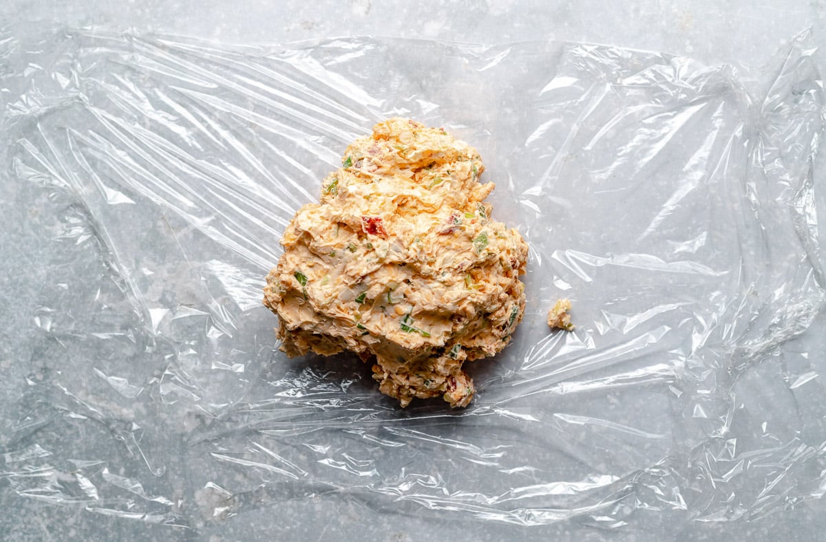 A Bacon Cheddar Cheese Ball mixture has been scooped atop a clear sheet of plastic wrap that sits atop a blue-gray textured surface.