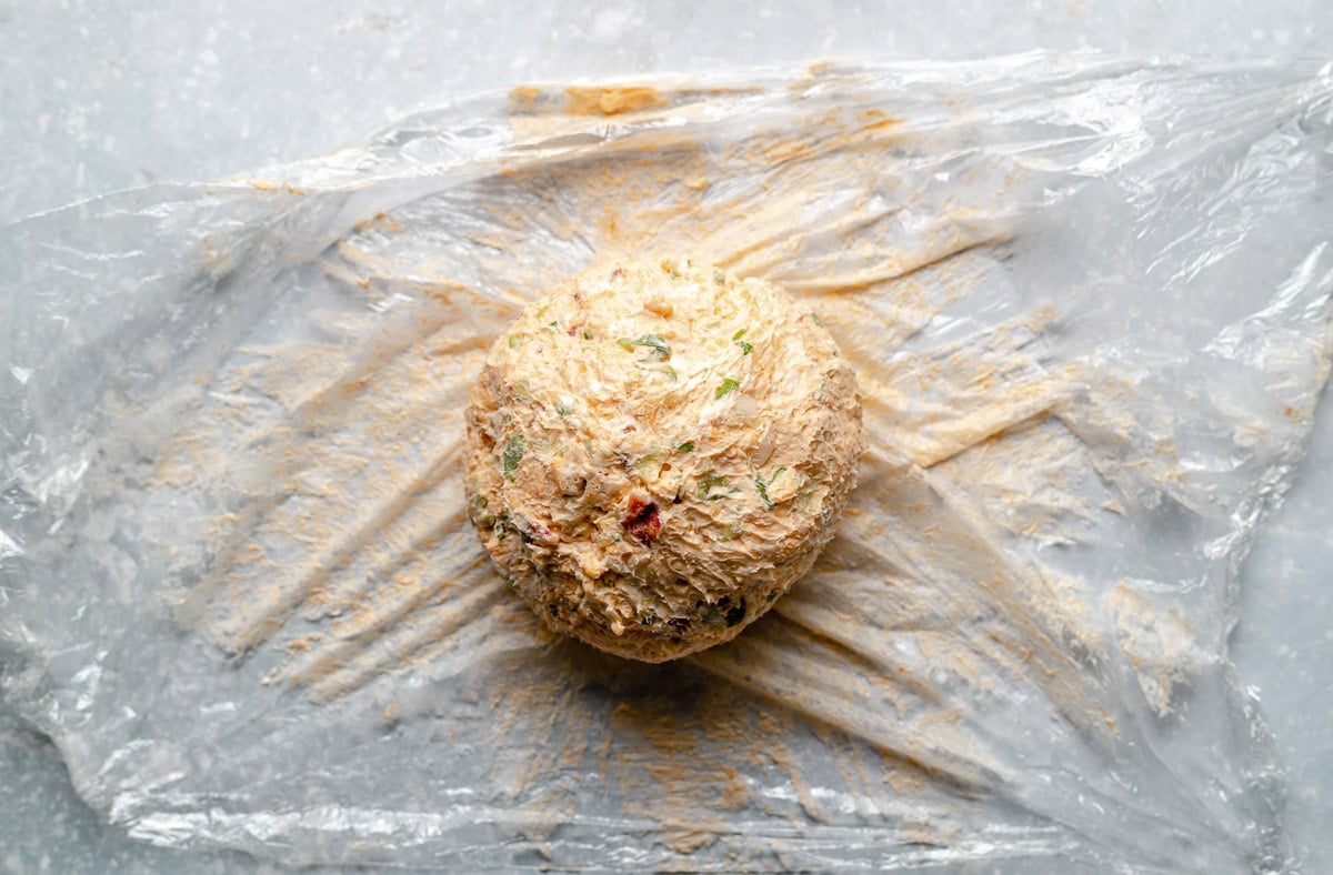 A chilled and formed cheese ball made with cream cheese and beer sits atop a piece of clear plastic wrap that has been unfolded revealing the set cheese ball ready to be coated. The plastic wrap and cheese ball sit atop a blue-gray textured surface.