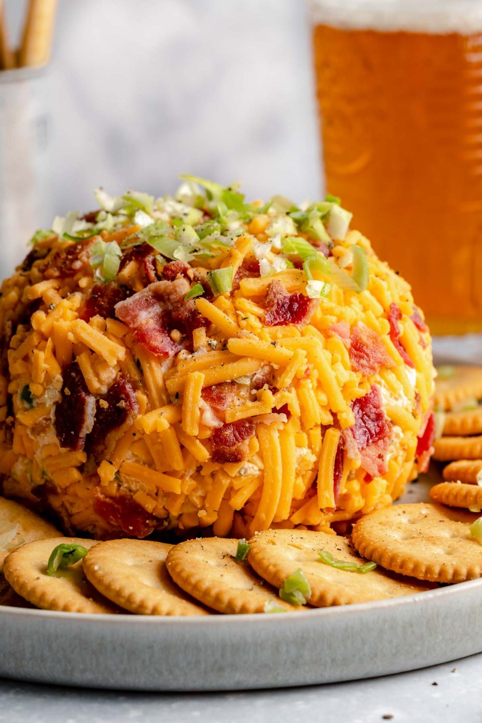 A side angle shot of a Bacon Cheddar Cheese ball sits atop a gray ceramic platter surrounded by a ring of Ritz crackers. The platter rests atop a gray-blue textured surface. A pint of beer and a silver container filled with breadstick crackers sits in the background out of focus.