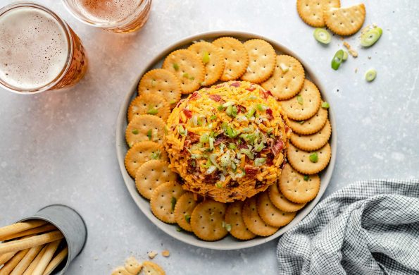An overhead shot of a Bacon Cheddar Cheese ball that sits atop a round gray ceramic platter surrounded by a complete ring of Ritz crackers that have bits of green onion, bacon and shredded cheddar cheese sprinkled on top. The platter rests atop a gray-blue textured surface. A couple of pints of amber-colored beer, a few Ritz crackers, green onions, a silver container filled with breadstick crackers, and a blue and white plaid linen napkin rest on the surface surrounding the platter.