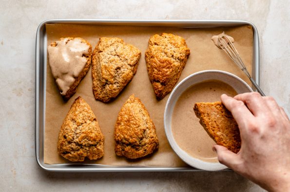 Dipping chai scones in maple glaze. Baked scones & bowl of glaze atop a parchment-lined baking sheet. A woman's hand dunks 1 of the scones into the glaze.