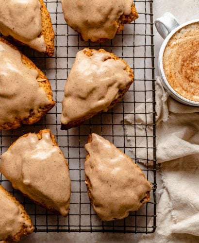 Glazed Chai Scones sit atop a metal baking rack. Next to the rack are a beige linen napkin, a whole cinnamon stick, & a mug with a chai latte in it.