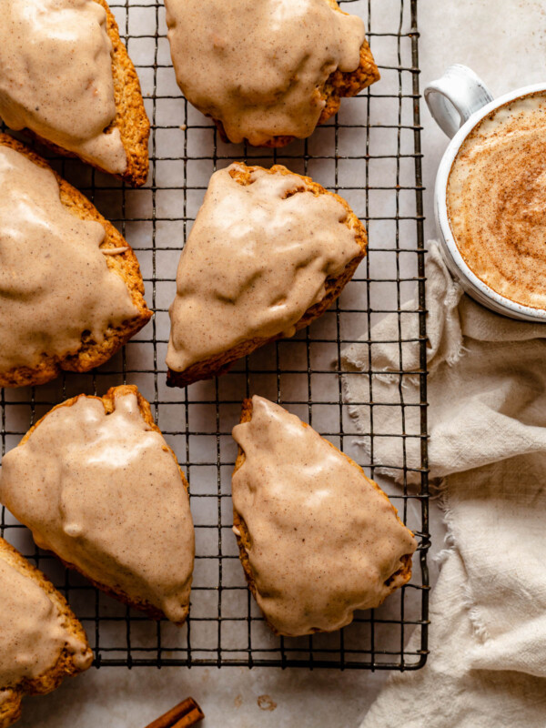 Glazed Chai Scones sit atop a metal baking rack. Next to the rack are a beige linen napkin, a whole cinnamon stick, & a mug with a chai latte in it.