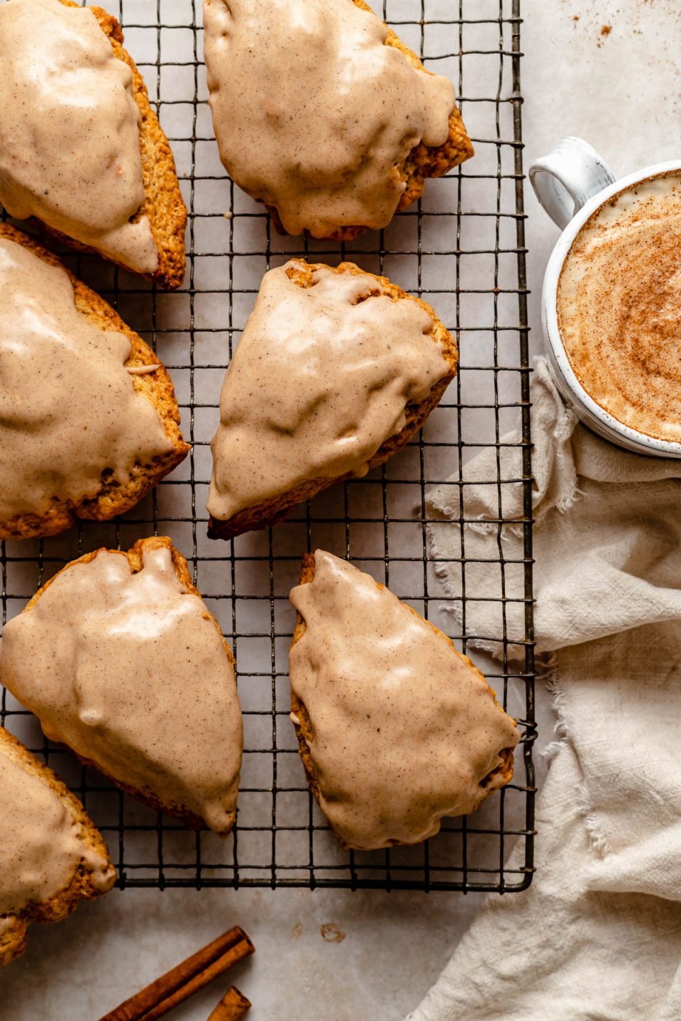 Glazed Chai Scones sit atop a metal baking rack. Next to the rack are a beige linen napkin, a whole cinnamon stick, & a mug with a chai latte in it.
